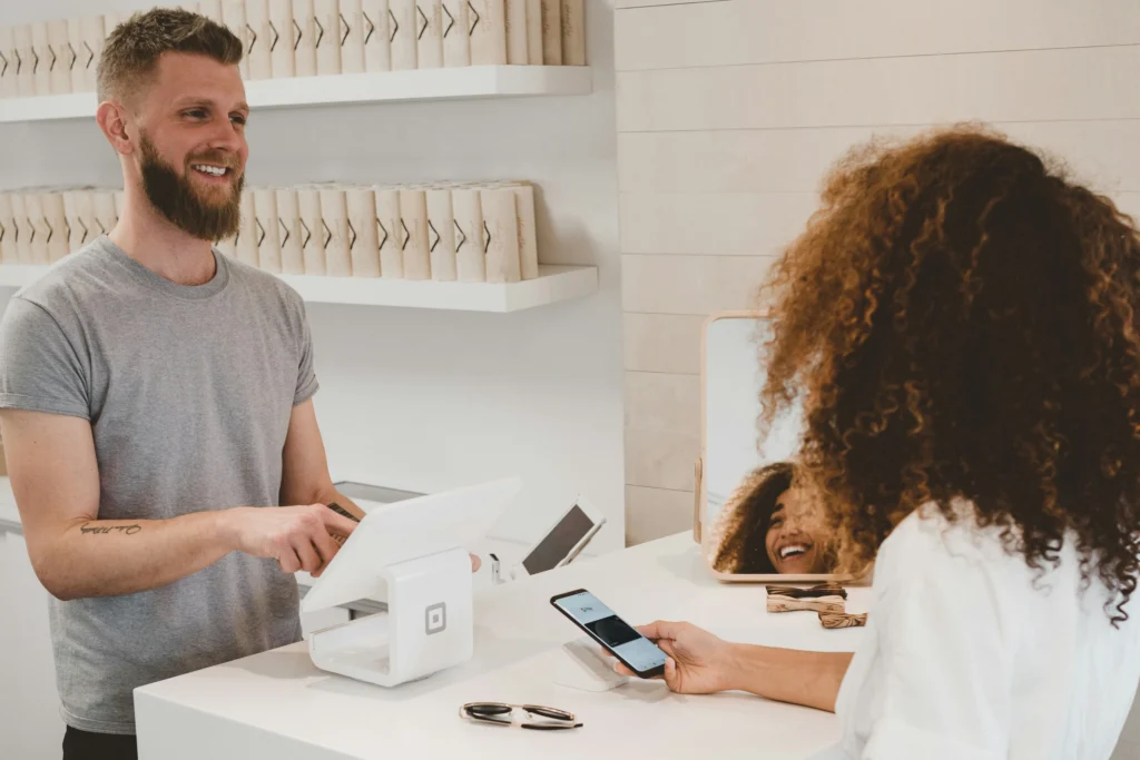 A man at a checkout counter, smiling while assisting a woman holding a smartphone for payment. The setting is modern, calm, with neatly arranged shelves.