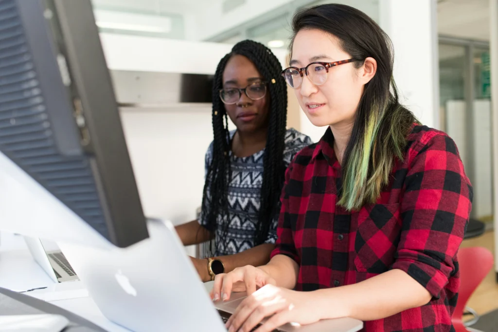 Two female professionals, one with green-streaked hair, collaborating over a laptop in a Canadian office, representing a focused workforce defining new future skills as part of an AI talent strategy.