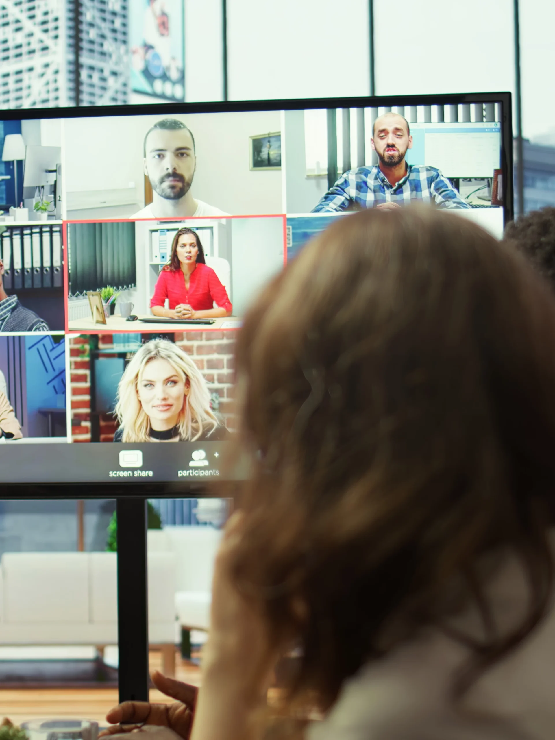A large screen displays a video conference with multiple remote participants while an in-person attendee listens during a hybrid office meeting.