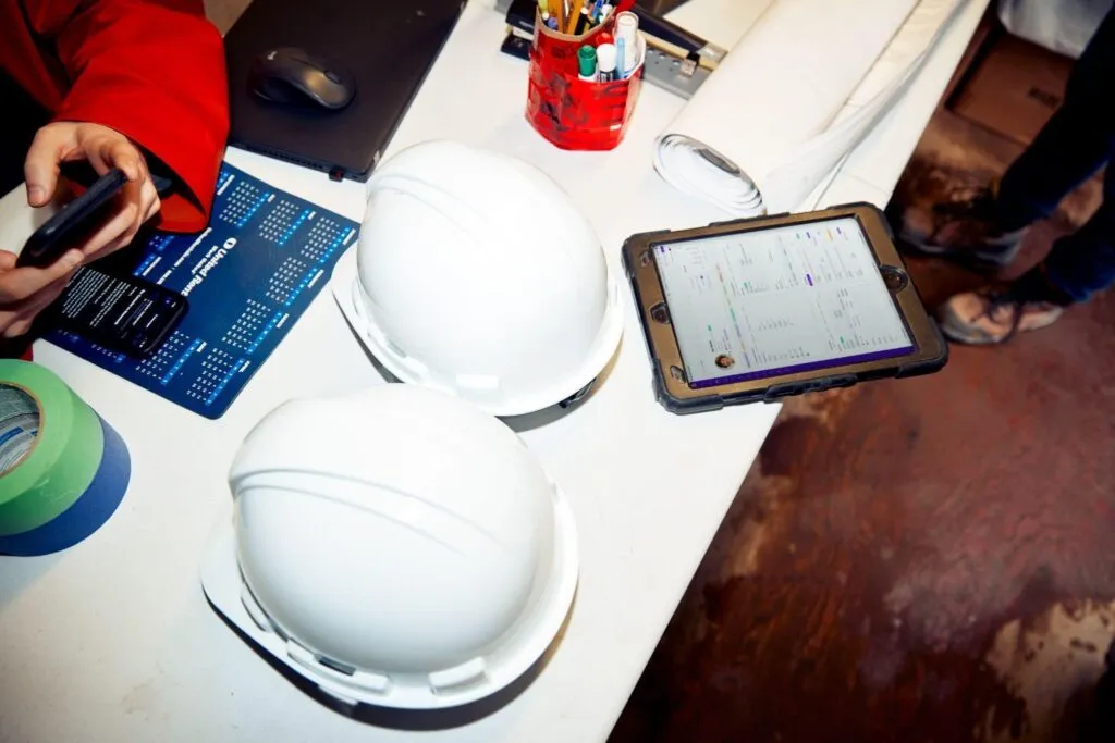 A rugged tablet displaying Employment Hero's HRIS software resting next to white hard hats on a construction site table, demonstrating how small businesses in Canada can effectively manage HR for deskless and site-based teams.