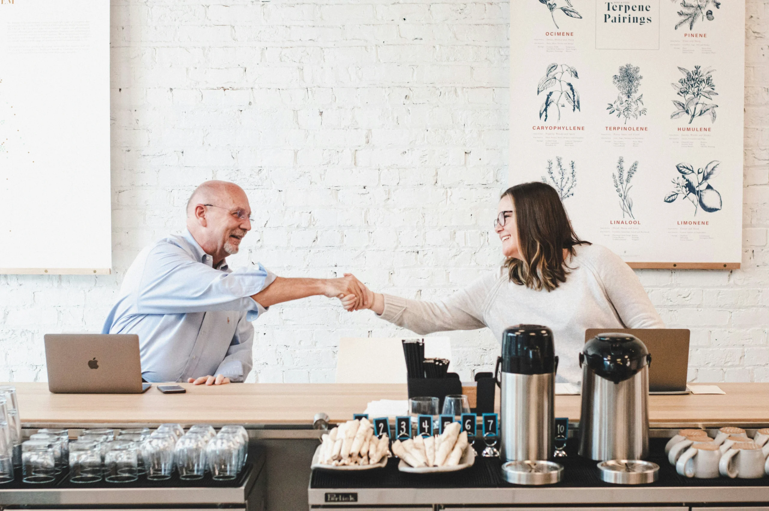 A man and woman smiling and shaking hands across a café counter, with laptops beside them. The mood is friendly and professional, against a backdrop of botanical illustrations.