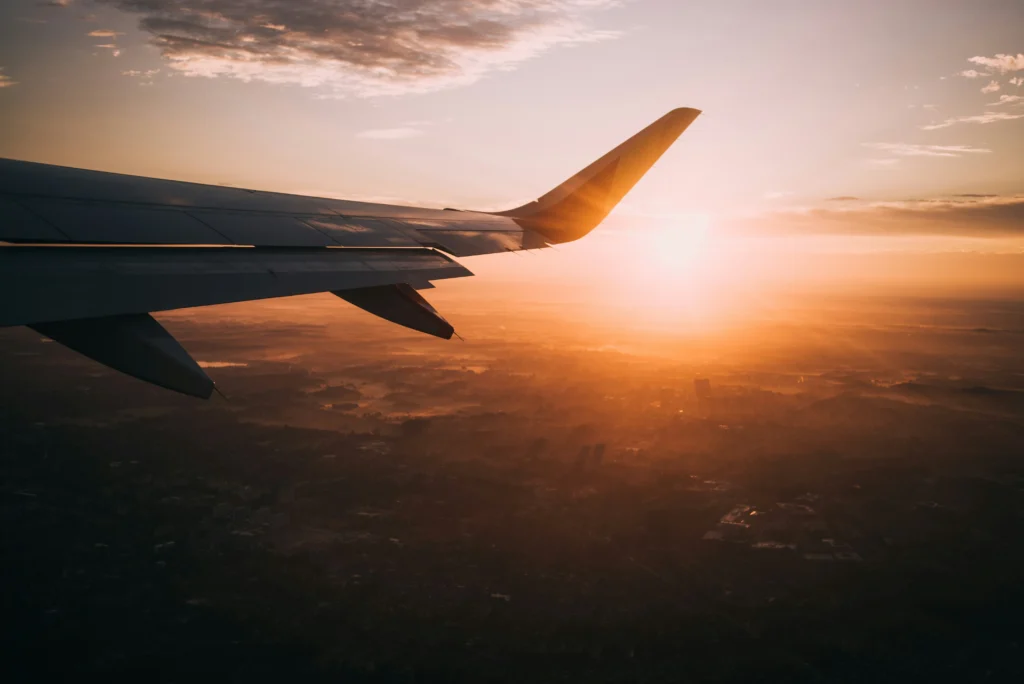 An airplane wing silhouetted against a vibrant orange sunset, viewed from a passenger window overlooking a hazy landscape.