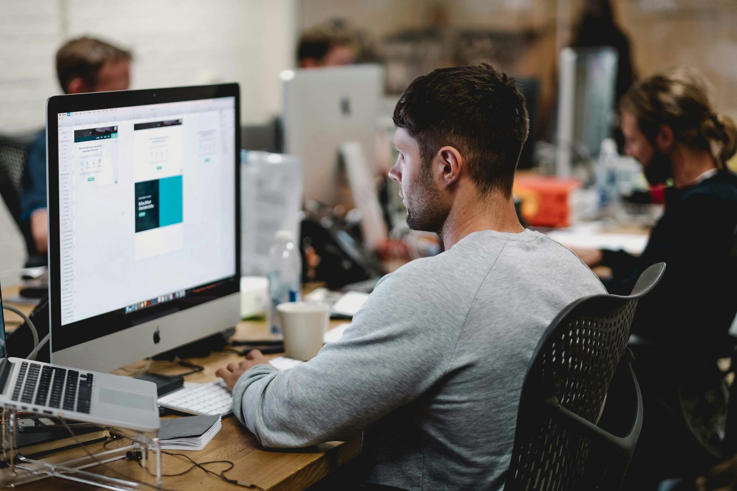 A man in a gray sweater works intently at a computer in a modern office. Other workers in the background focus on their screens. The atmosphere is focused and collaborative.