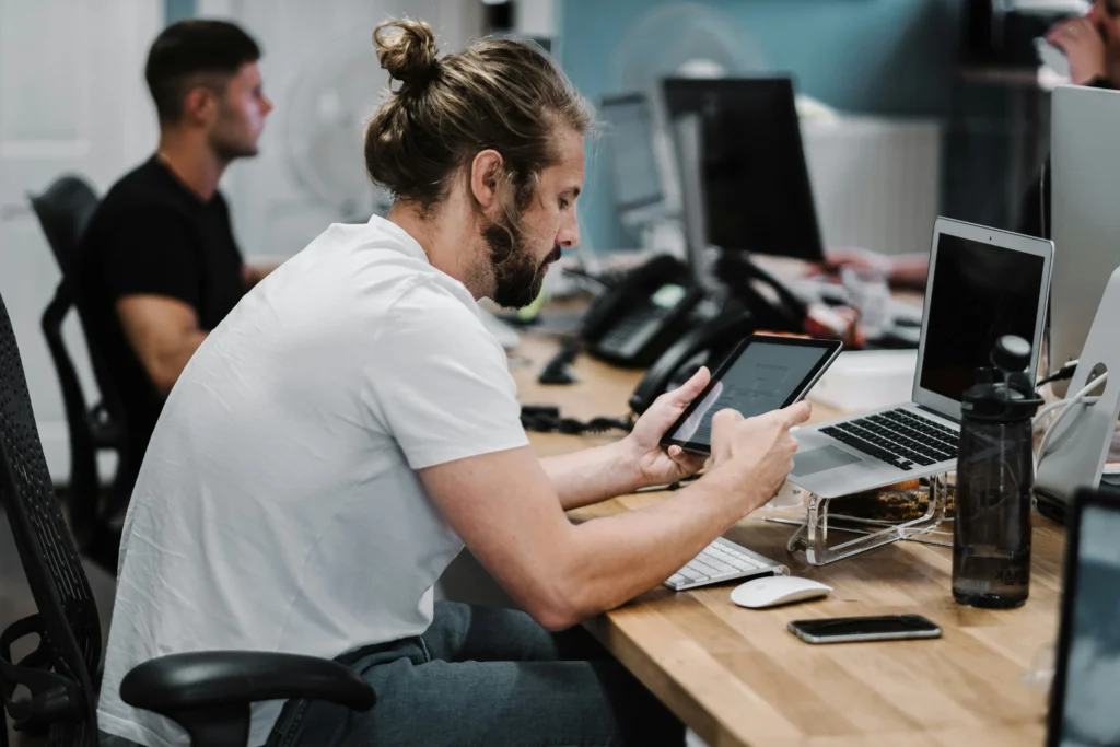 A man with a bun and beard focuses on a tablet at a wooden desk with a laptop, smartphone, and water bottle; another person works in the background.