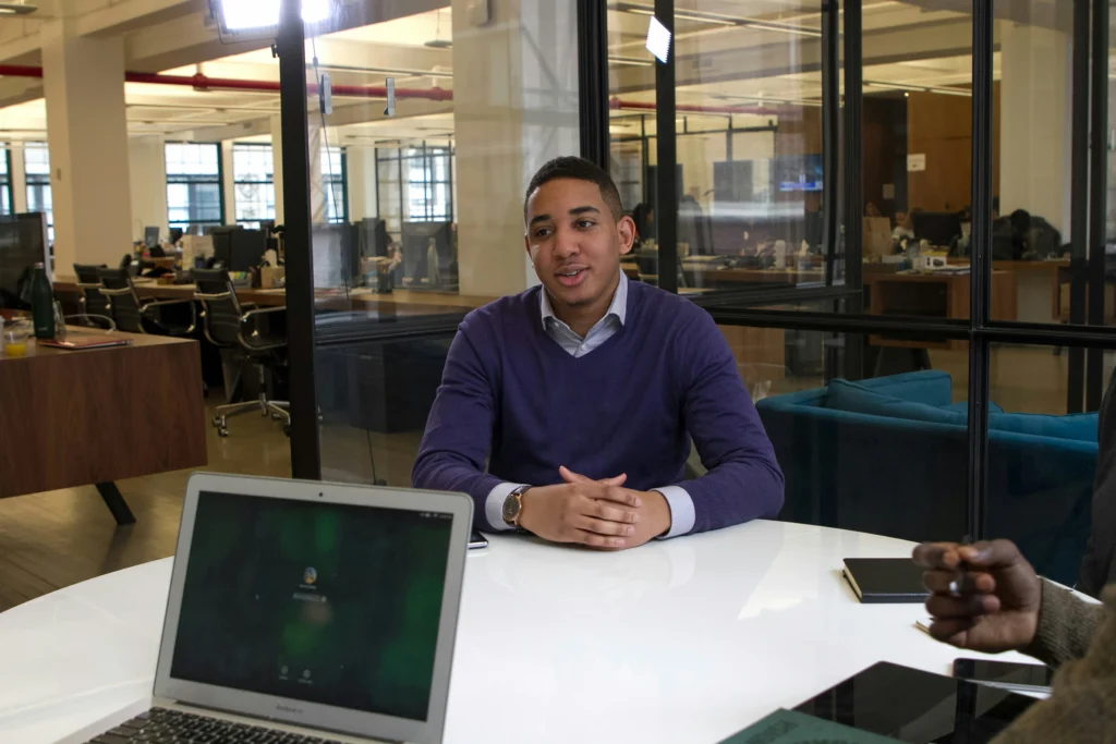 A person in a purple sweater sits at a white table in a modern office, engaged in conversation. A laptop and notebook are visible on the table.