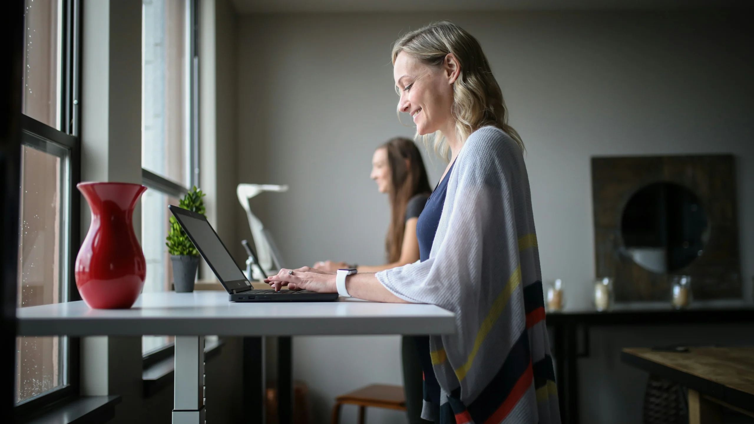 A woman sits at a desk, focused on her laptop, with a notepad and pen beside her.
