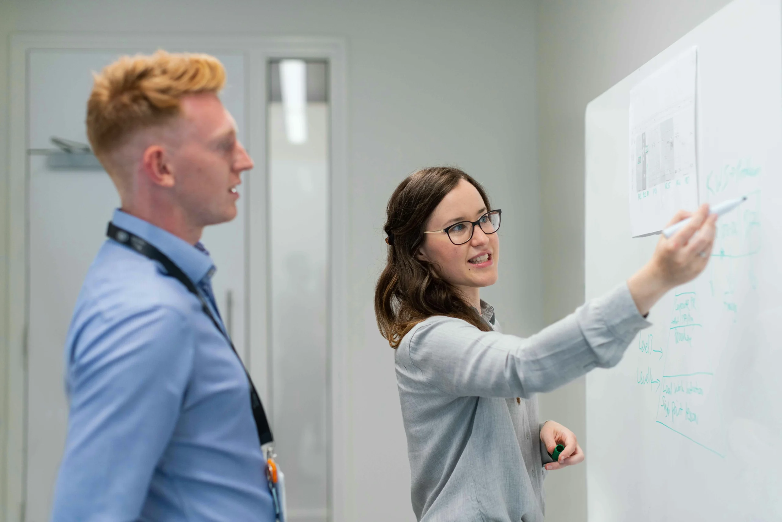A woman smiles while explaining a chart on a whiteboard to a man in an office. The atmosphere is collaborative and focused, with notes in green marker.