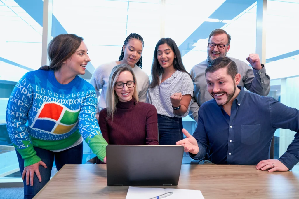 A diverse group of six people stand around a laptop, smiling and expressing excitement in a modern office setting, conveying teamwork and success.