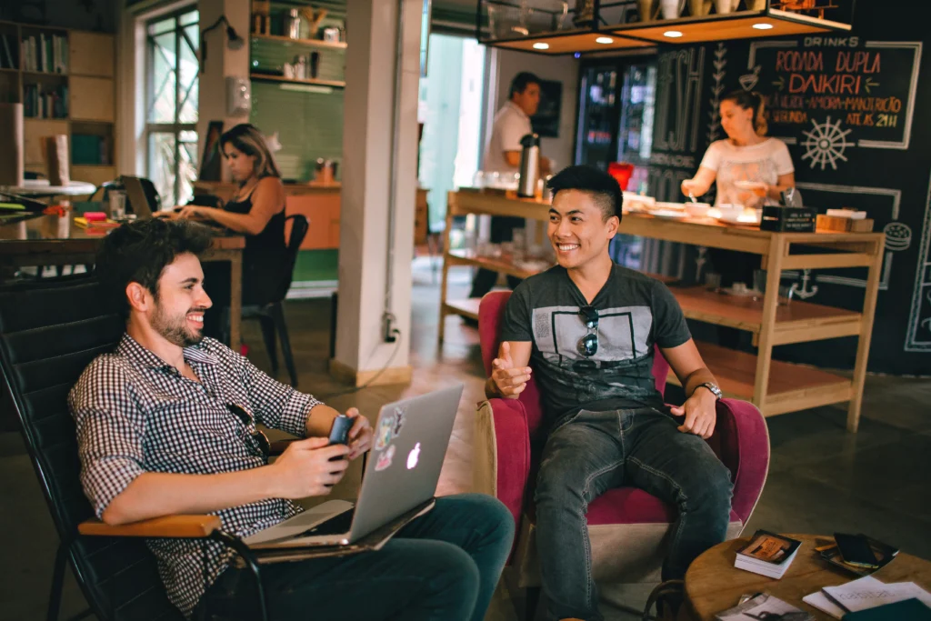 Two male professionals sitting in relaxed chairs in a casual workspace, laughing and discussing something on a laptop, illustrating how workers can collaborate and adapt to new technologies amid AI job displacement.