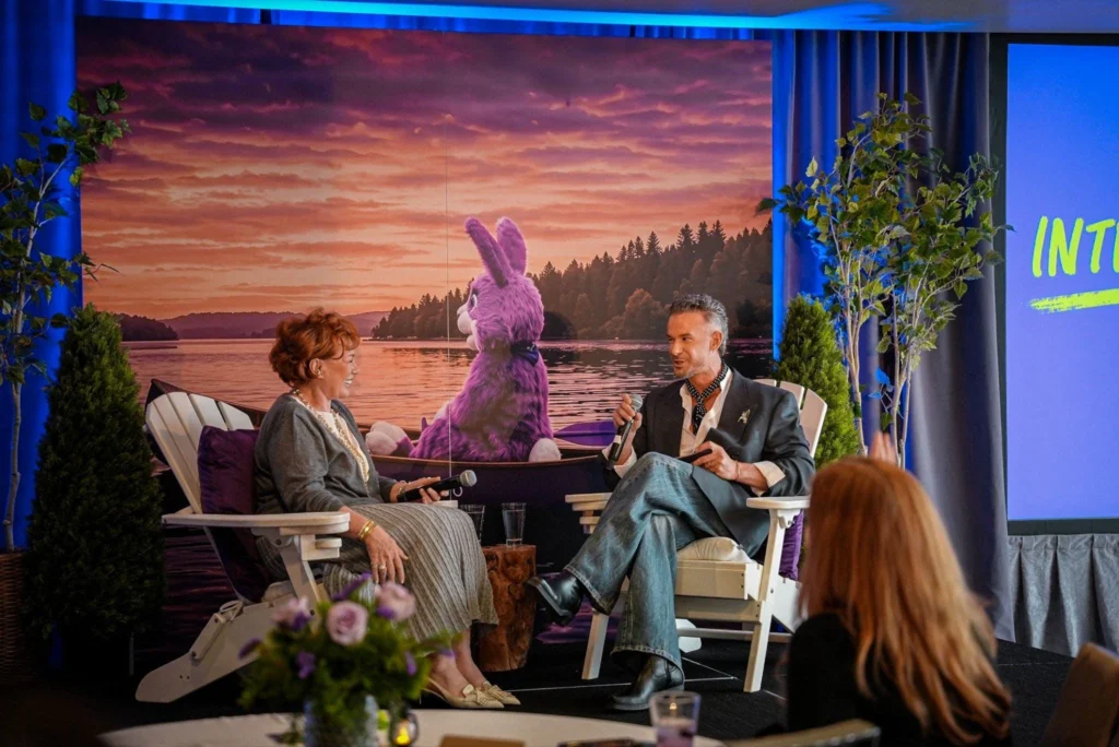 Two people are seated in white chairs on stage, engaged in conversation with microphones. A backdrop features a purple rabbit overlooking a lake at sunset.