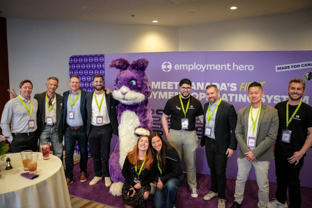 A group of smiling people, including a person in a purple and white furry mascot costume, pose for a photo against a purple banner promoting Employment Hero.