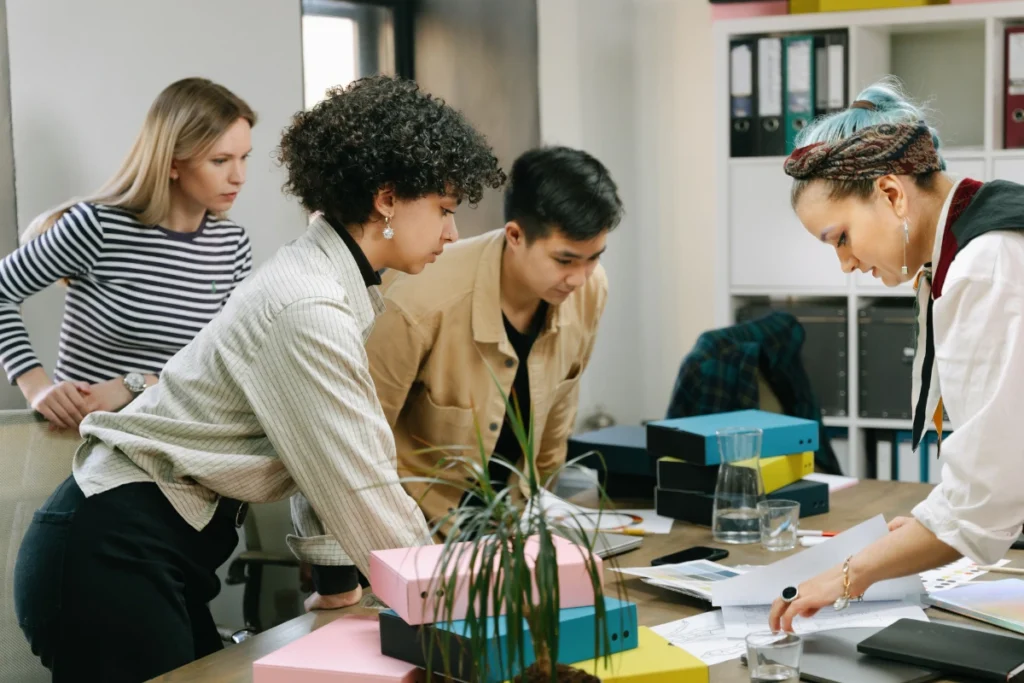 Four people engaged in a focused discussion around a cluttered meeting table, examining documents. Office shelves with colorful folders in the background.