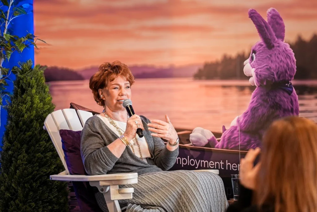 A woman with a microphone sits on a white chair, speaking, against a sunset backdrop featuring a purple cartoon rabbit by a lake, conveying a relaxed atmosphere.