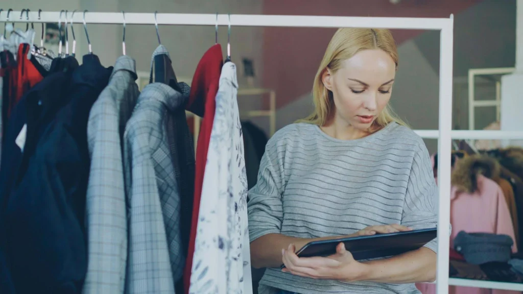 A woman with blonde hair in a gray striped sweater looks at a tablet while standing beside a clothing rack filled with various garments in a store.