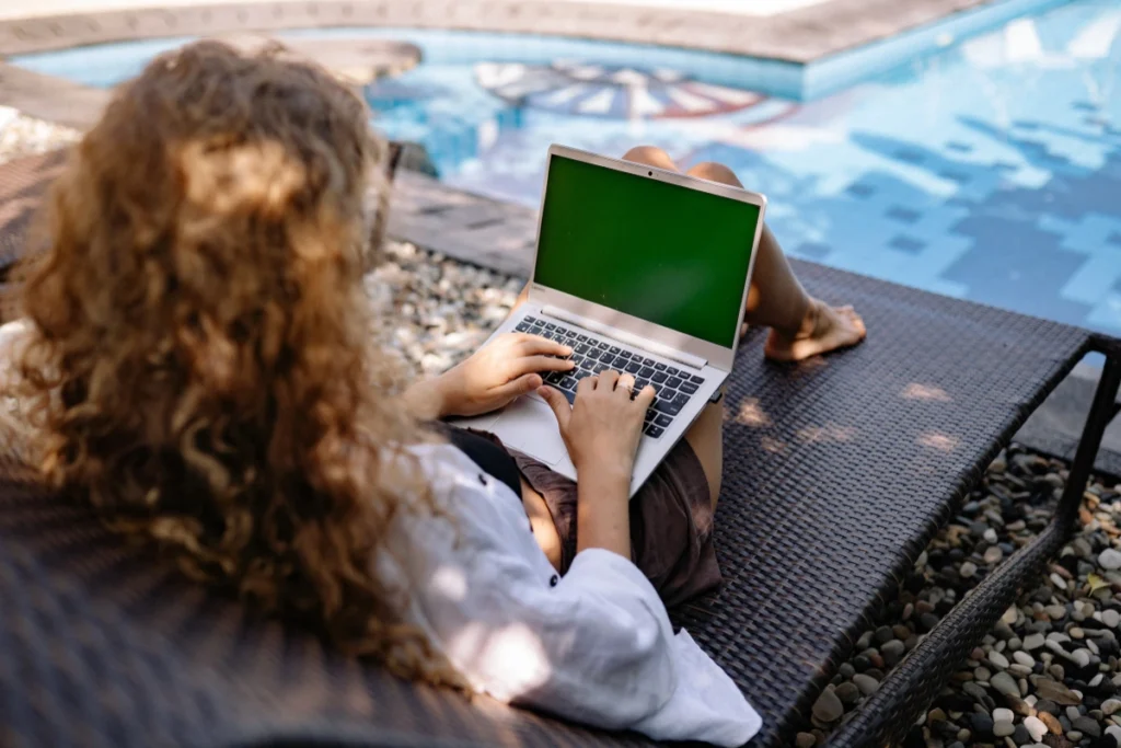 A person with curly hair works on a laptop with a green screen while lounging by a pool. The scene conveys relaxation and remote work.