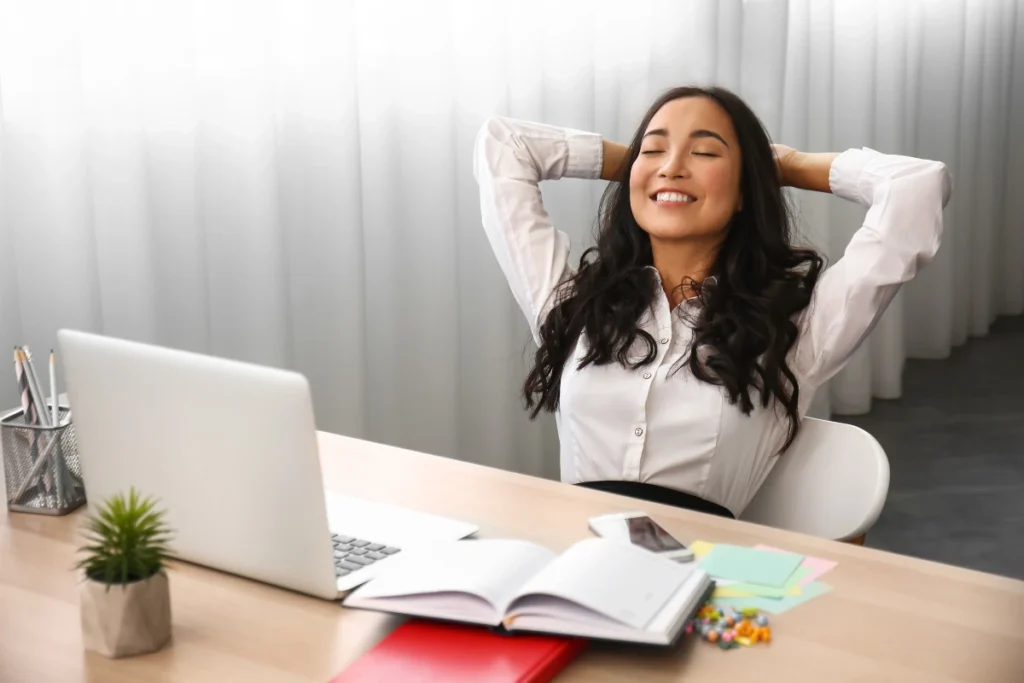 A relaxed woman sits at a desk with a laptop and open notebook, smiling with eyes closed and hands behind her head. The scene conveys a sense of calm and contentment.