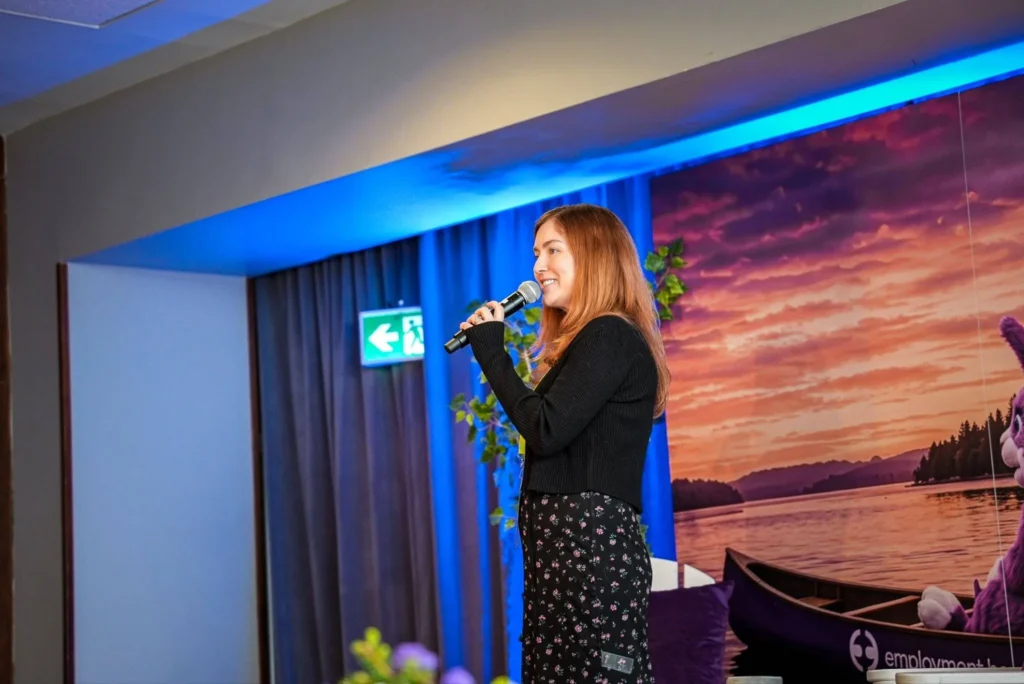 A woman in a black sweater and floral skirt speaks into a microphone on stage. Behind her is a vibrant sunset lake scene, creating a warm atmosphere.