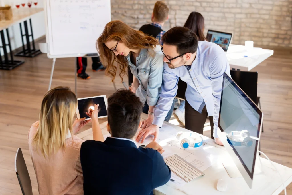 A group of colleagues collaborates energetically around a table with computers and papers, focused on shared tasks. The setting is a modern office.