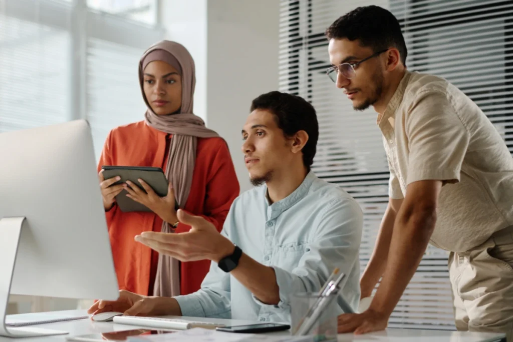 Three colleagues collaborate at a desk, focused on a computer screen. One points while another takes notes on a tablet, creating an engaged atmosphere.