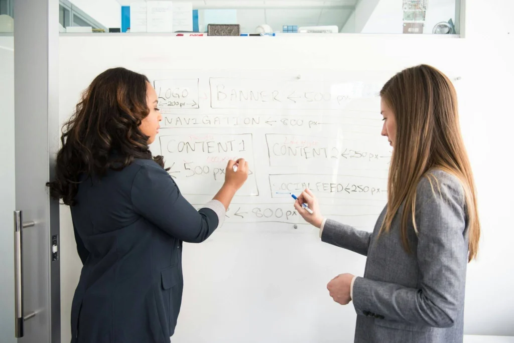 Two women collaborate at a whiteboard in an office, writing and drawing diagrams. They appear focused and engaged in a planning session.