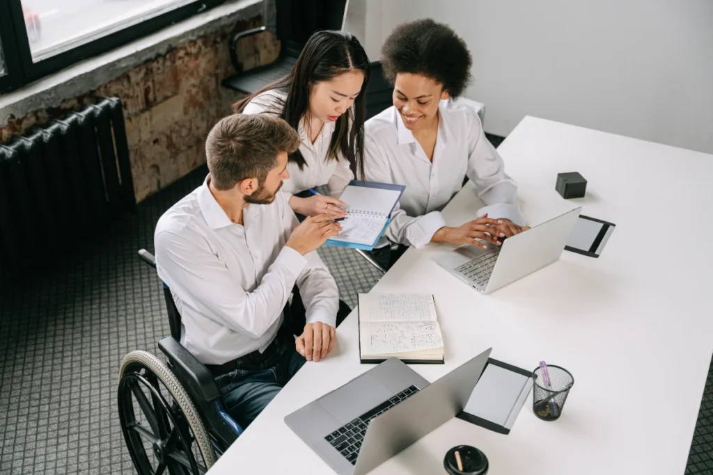 Three colleagues collaborate around a white table with laptops and notebooks. They are smiling and discussing a document in an office setting.