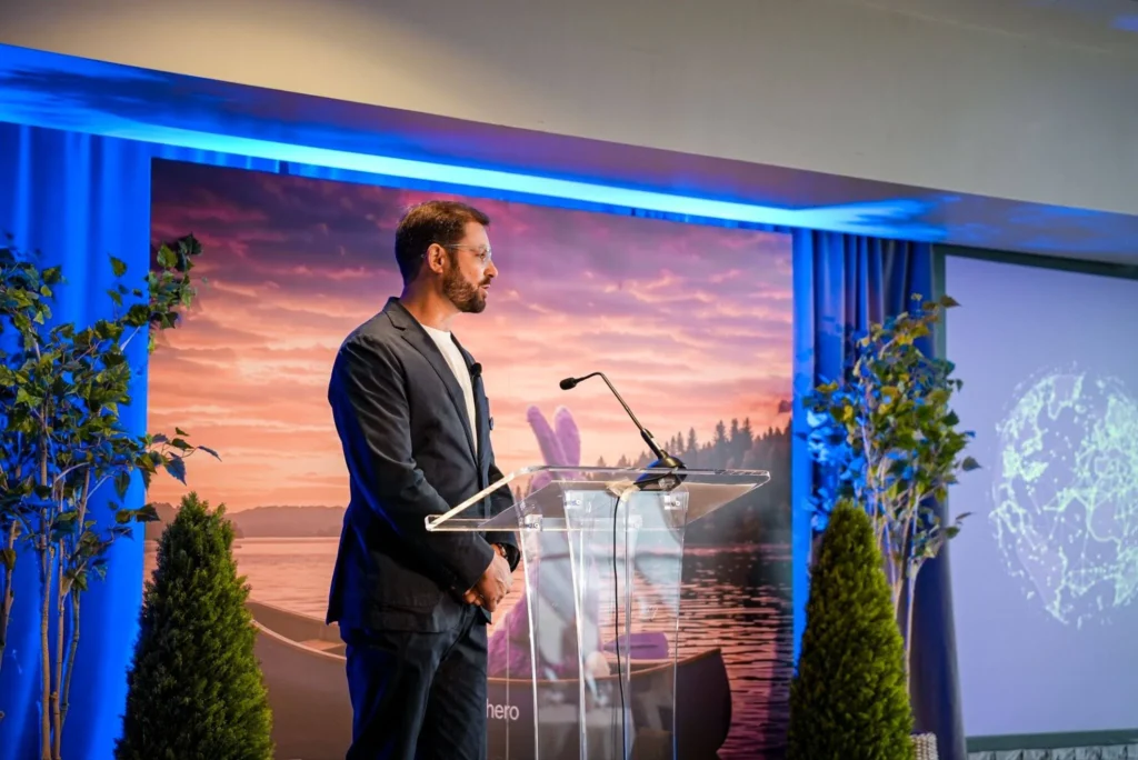 A man in a suit stands at a clear podium on a stage, speaking with a sunset lake backdrop and digital globe screen. The setting is professional and serene.