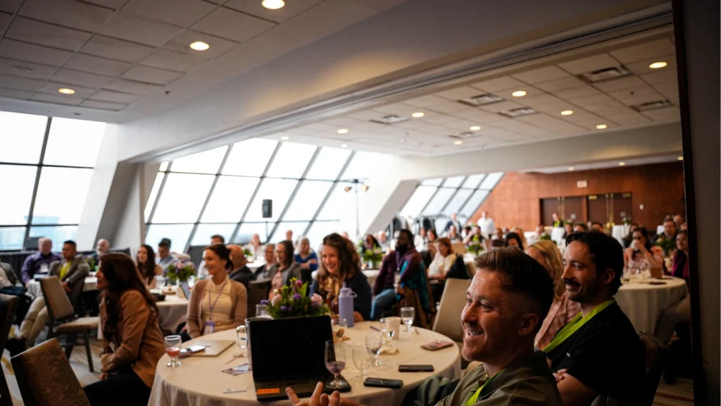 A conference room filled with smiling attendees seated at round tables. Large windows and dim lighting create a warm, engaging atmosphere.