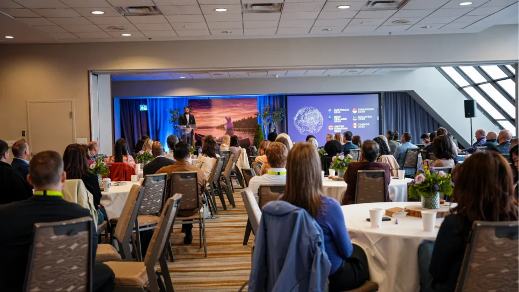 Conference room filled with attendees seated at round tables, facing a stage where a presenter stands at a podium. A large screen displays a digital globe.