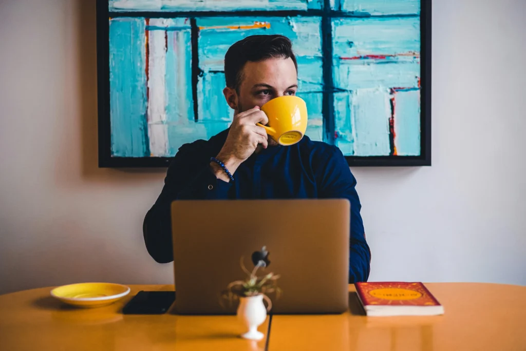 A person in a black shirt sits at a desk with a laptop, holding a yellow mug. A colorful abstract painting hangs in the background. The scene feels contemplative.