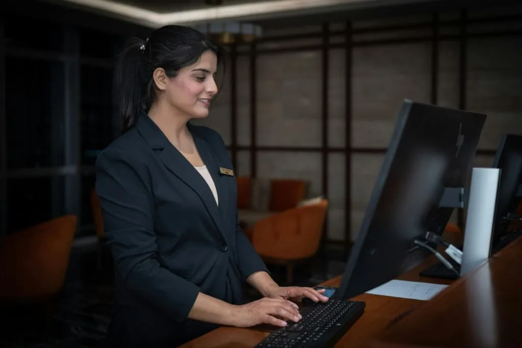 A smiling female hotel receptionist in a dark blazer typing at a front desk computer, illustrating the dedicated hospitality talent in Canada who benefit from a clear career progression plan to prevent burnout.