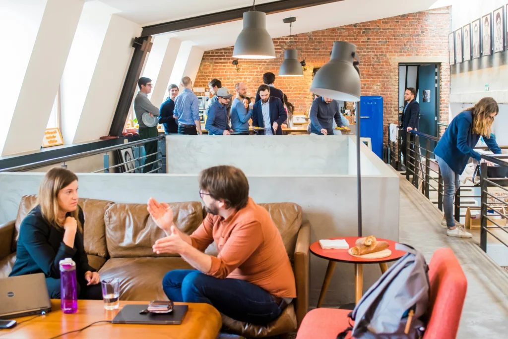 A lively office space with people engaging in conversation. In the foreground, a man and woman chat on a sofa. Others gather near a brick wall, creating a collaborative atmosphere.