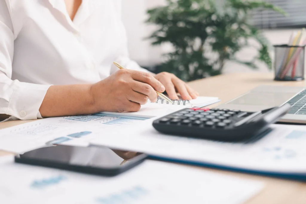A person in a white shirt writes in a notebook at a desk with charts, a calculator, and a laptop, conveying a focused, professional tone.