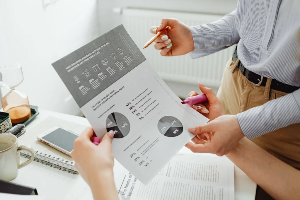 Two people analyzing a printed report with pie charts, holding pens in a bright office. A phone and coffee mug rest on the desk, indicating a professional setting.