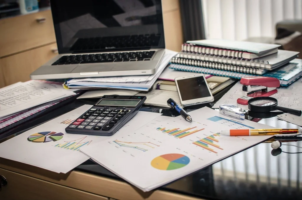 A cluttered desk with a laptop, calculator, smartphone, charts, notebooks, and stationery. The scene conveys busyness and productivity.