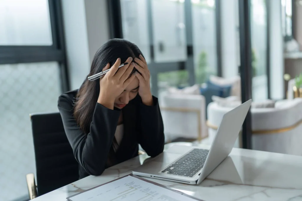 A woman sits at a desk with her head in her hands, looking stressed. A laptop and documents are in front of her in a bright, modern office space.