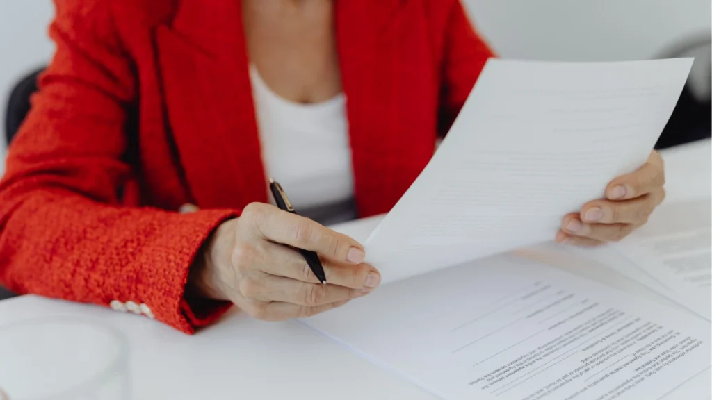 A person in a red blazer reviews a document at a white desk, holding a pen. Papers are spread out, suggesting a professional or focused setting.