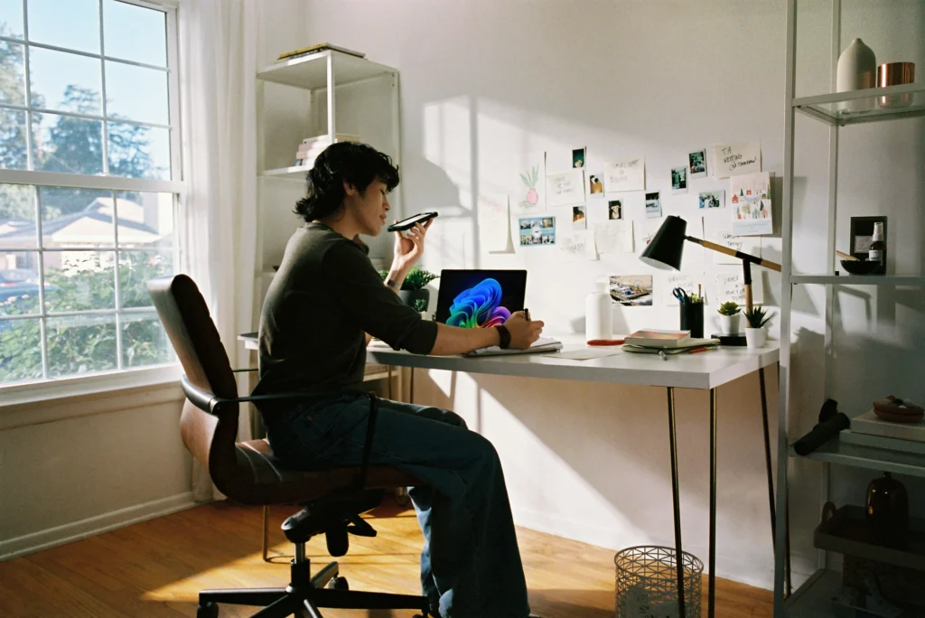 A person sits at a desk in a sunlit room, speaking into a smartphone. A laptop with a colorful screen is open, surrounded by notes and plants, suggesting productivity and focus.