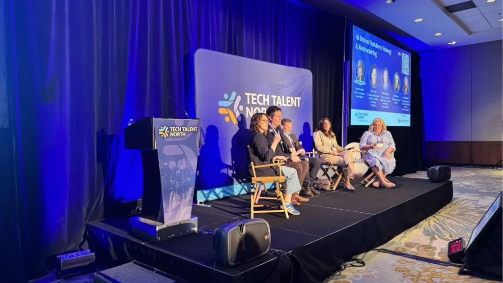 A panel of four people sits on stage at the Tech Talent North conference. The speaker is talking into a microphone. A large screen shows panel details.