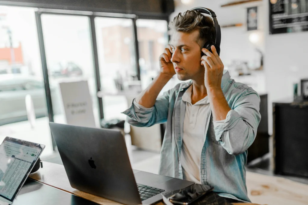 A young man in a casual shirt adjusts his headphones while seated at a café table with a laptop and tablet. He appears focused, suggesting he is working or studying.