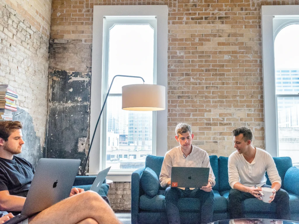 Three people work collaboratively in a modern brick-walled office, using laptops. Natural light filters in through tall windows, creating a focused yet relaxed atmosphere.