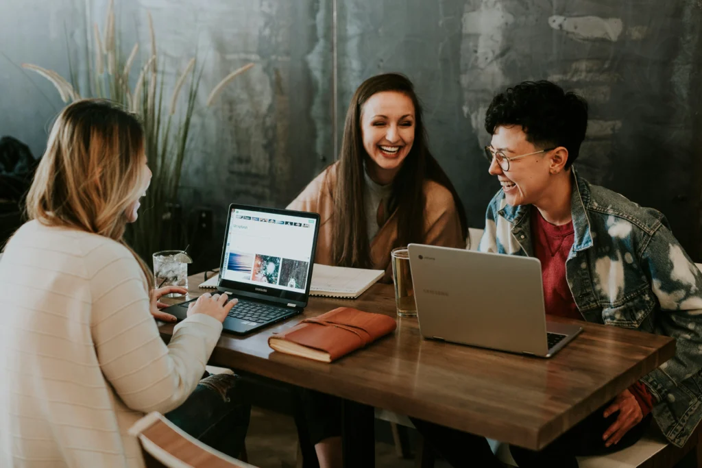 Three young adults laughing and working together with laptops at a wooden table in a cafe.