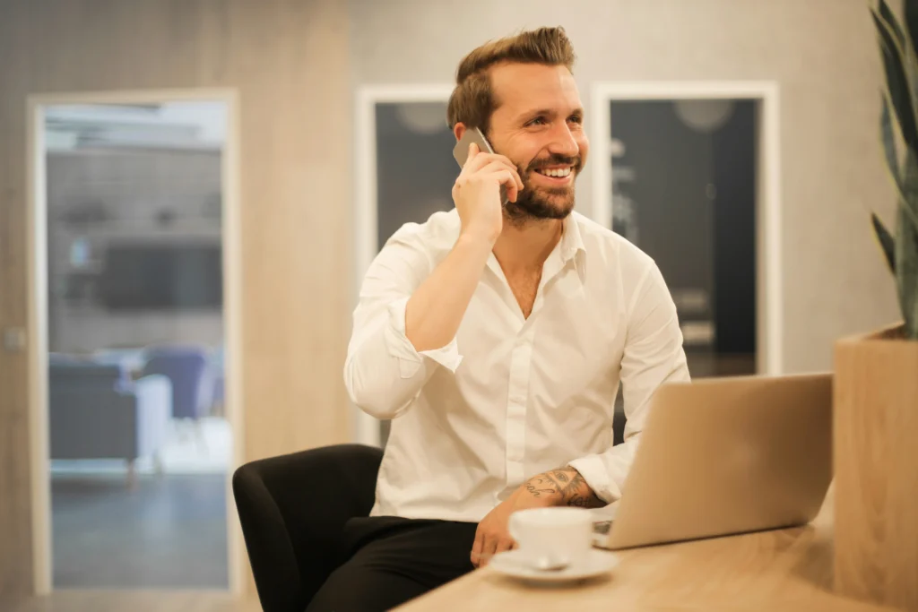 Smiling man sitting at a laptop, talking on his smartphone in a modern, brightly lit office environment.