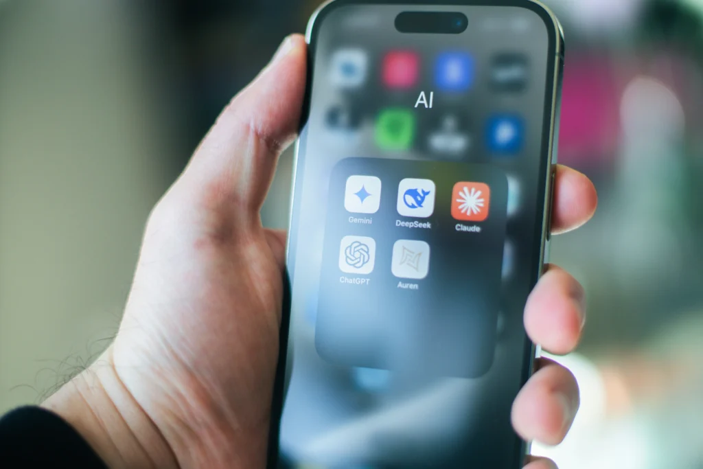 Close-up of a hand holding a smartphone displaying an AI folder with apps: Gemini, DeepSeek, Claude, ChatGPT, and Auren. Modern tech context.
