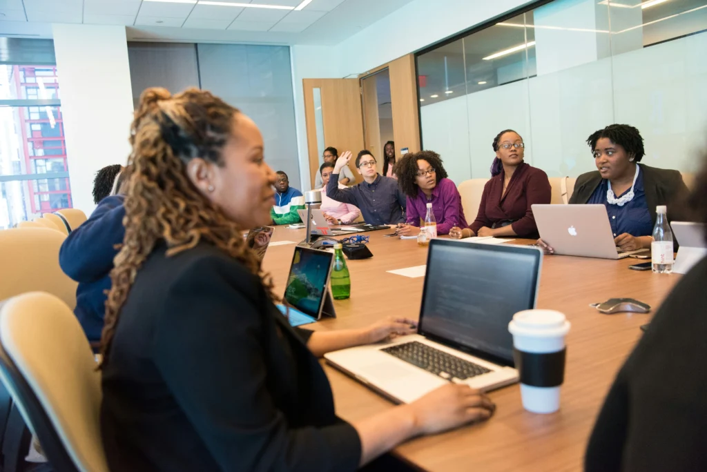 A diverse group of people in a bright meeting room, engaged in discussion. Laptops and notebooks are on the table, conveying a collaborative atmosphere.