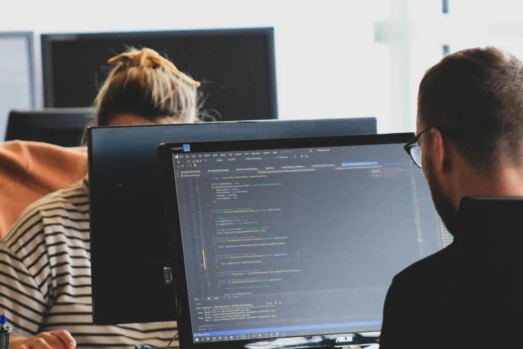 Two people work at desks with computer screens displaying code. A striped shirt is visible in the background, suggesting a collaborative office environment.