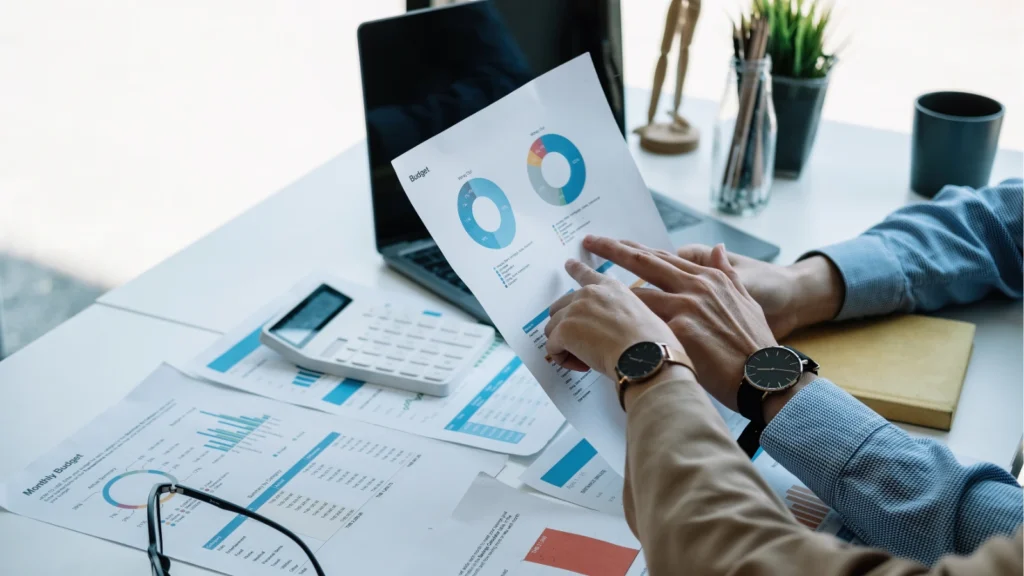 Two professionals analyze financial charts and graphs on paper at a desk. A laptop, calculator, and potted plant are visible, conveying a focused work setting.