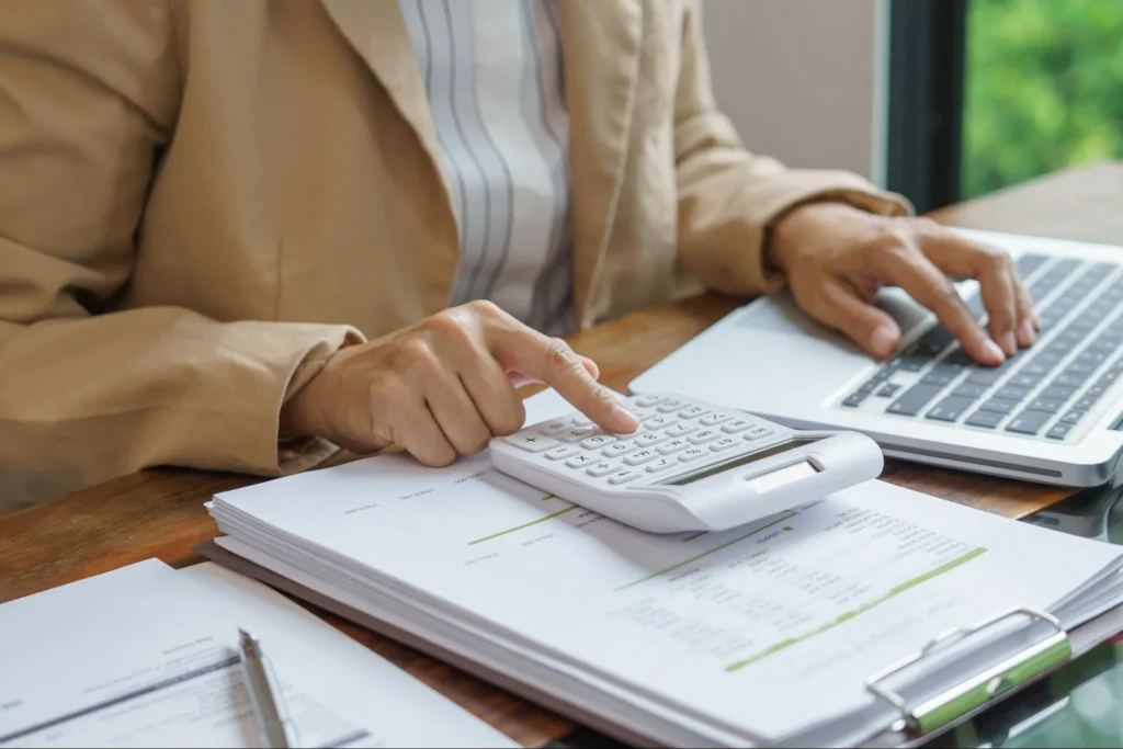 Close-up of a person in a beige blazer using a white calculator and a laptop to work on financial documents at a wooden desk.