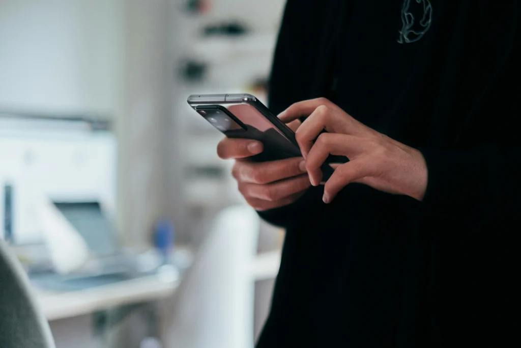 A person in dark clothing holding and typing on a modern smartphone with a large camera module, with a blurred office background.