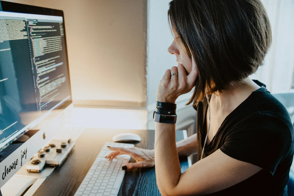 A woman with short brown hair is intently looking at code on a computer screen. She sits at a desk with thoughtful focus, conveying concentration and dedication.