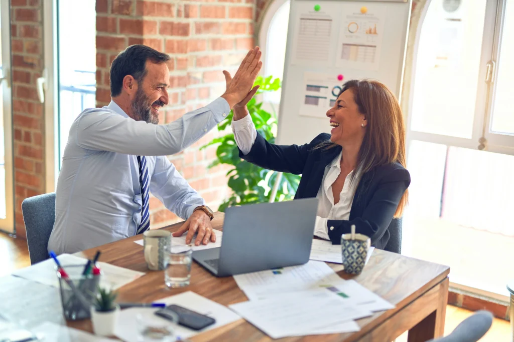 A man and woman in a business setting cheerfully share a high-five over a wooden table with laptops and papers, conveying success and teamwork.
